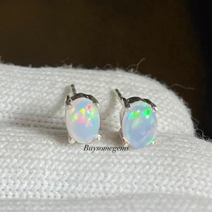 Pode incluir: Um par de brincos de opala ovais com pinos de prata. As opalas exibem um jogo de cores com flashes de verde, rosa e amarelo. O texto "Buysomegems" é visível.