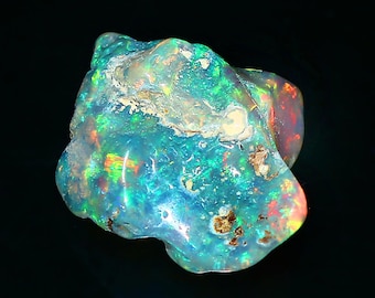 May include: A vibrant, uncut opal gemstone with a mix of colors. The stone displays a play of color with flashes of green, blue, red, and yellow. The opal has a rough, natural texture and is set against a dark background.