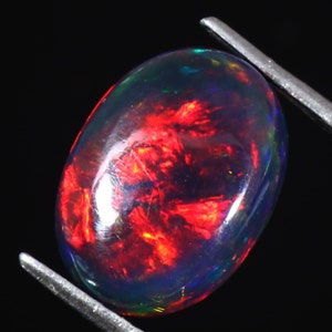 Pu&ograve; includere: Una pietra preziosa di opale nero di forma ovale scura con un gioco di colori rosso e verde iridescente.