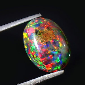 Könnte beinhalten: Ein lebendiger schwarzer Opal-Edelstein mit einem farbenfrohen Spiel aus roten, grünen, blauen und gelben Blitzen. Der ovalförmige Edelstein ist in einer klaren, polierten Fassung gefasst.
