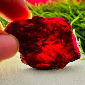 May include: A large, deep red, rough-cut gemstone with a translucent appearance. The gemstone has a natural, irregular shape and is likely a ruby.