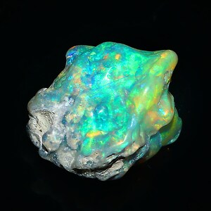 May include: A vibrant, raw opal gemstone with a rough, textured surface. The opal displays a striking play of color, with flashes of green, blue, and yellow. The stone has a natural, irregular shape, and a gray base.