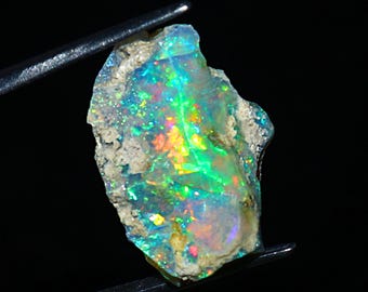 May include: A rough opal gemstone held by tweezers. The opal displays a vibrant play-of-color, with flashes of green, blue, yellow, and red. The stone has a raw, natural appearance with a textured surface and a light-colored matrix.