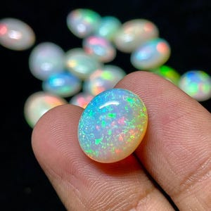 Op de afbeelding: Een close-up van een enkele ovale opaal edelsteen met een regenboog van kleuren, waaronder groen, blauw en rood. De opaal is iriserend en heeft een witte basis.