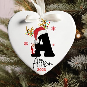 May include: White heart-shaped Christmas ornament with a black letter A, a Santa Claus peeking out from behind the letter, and the name Allison and the year 2023 printed below.
