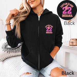 May include: Black zip-up hoodie with the text "Caitlin Clark 22 Indiana" in pink. The back of the hoodie also features the same design in a circular format. The hoodie has a front pocket and a zipper closure.
