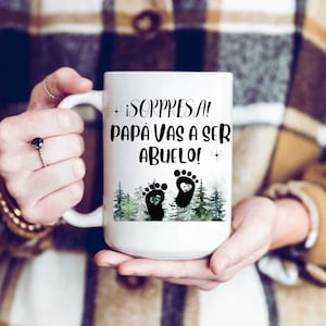 May include: White ceramic mug with a green watercolor forest scene and two black baby footprints with hearts. The text on the mug reads "¡SORPRESA! PAPÁ VAS A SER ABUELO!"