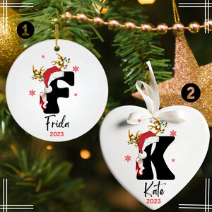 May include: Two white ceramic Christmas ornaments, one round and one heart-shaped. The round ornament features a black letter "F" with a Santa hat and reindeer antlers. The text "Frida 2023" is below the letter. The heart-shaped ornament features a black letter "K" with a Santa hat and reindeer antlers. The text "Kate 2023" is below the letter.