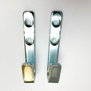 May include: Two silver-colored metal wall hooks with a polished finish. Each hook features a flat, elongated vertical mounting plate with two circular, countersunk screw holes positioned one above the other, and a single upward-curving hook at the base.