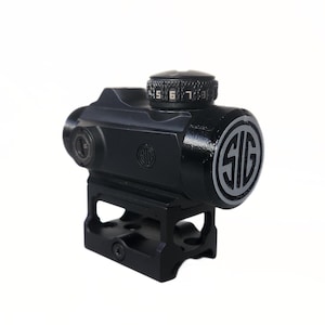 Op de afbeelding: Een zwarte red dot sight met een matte afwerking. De sight heeft een cirkelvormige instelknop met genummerde instellingen en het "SIG" logo in een zilveren omtrek. De sight is gemonteerd op een zwarte basis.