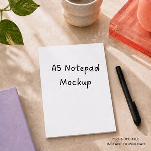 May include: A white A5 notepad mockup with the text A5 Notepad Mockup printed in black, accompanied by a black pen. The notepad is positioned vertically, featuring a clean surface for design display, suitable for digital stationery presentation.