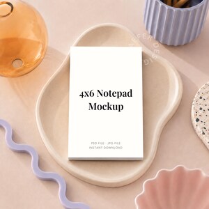 May include: A white 4x6 Notepad Mockup sits on a beige ceramic tray. The notepad has the text "4x6 Notepad Mockup" in black. Other decorative items include a light purple wavy object, a light blue ribbed container, and a pink bowl.