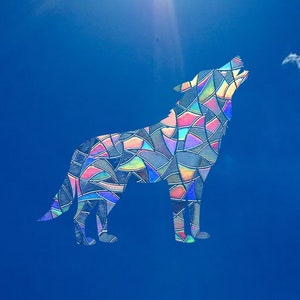 Könnte beinhalten: Eine farbenfrohe Wolfssilhouette im Glasmalerei-Stil vor einem leuchtend blauen Himmel. Der Wolf heult, und seine Form ist mit geometrischen Formen in Rosa, Blau, Grün und Gelb gefüllt. Einige weiße Wolken sind sichtbar.