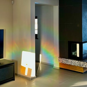 May include: A rainbow prism effect casts a colorful light pattern on a white wall in a modern living room. The light shines through a doorway and onto a white wall, creating a rainbow effect.