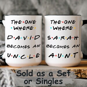 Op de afbeelding: Twee witte keramische mokken met zwarte handvatten en randen. Elke mok heeft tekst in het zwart, met gekleurde stippen. Eén mok zegt "THE ONE WHERE DAVID BECOMES AN UNCLE". De andere zegt "THE ONE WHERE SARAH BECOMES AN AUNT".