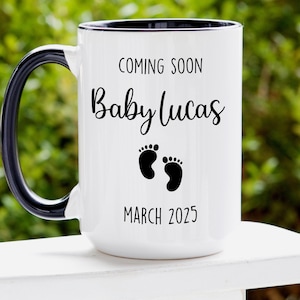 May include: White ceramic mug with a black handle and rim. The mug has black text that reads "Coming Soon Baby Lucas March 2025" and two black footprints.