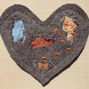 May include: A heart-shaped textile artwork in grey, featuring patches of blue, orange, and tan fabrics. These patches are stitched onto the grey base with orange thread, giving a textured, handcrafted look. The piece has a homespun, rustic aesthetic, with a unique design.