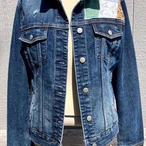 May include: A distressed denim jacket with a patchwork design on the shoulder. The jacket features silver buttons, two chest pockets, and a classic collar. The patchwork includes green, white lace, and patterned fabrics.