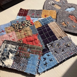 May include: A collection of square fabric coasters and a heart-shaped piece, all featuring patchwork designs made from various textile scraps. The pieces are joined with visible hand-stitched embroidery in contrasting colors like red and blue.