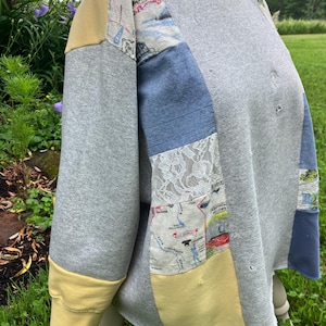 May include: Gray sweatshirt with patchwork details. The sleeves feature sections of blue denim, cream lace, and map fabric, accented with yellow cuffs and shoulder panels. The garment has a relaxed fit and visible stitching.