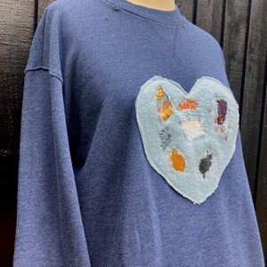 Könnte beinhalten: Ein marineblaues Sweatshirt mit einem hellblauen herzförmigen Aufnäher. Der Aufnäher ist mit verschiedenen bunten Stickereien verziert. Das Sweatshirt hat einen Rundhalsausschnitt und lange Ärmel. Der Stoff wirkt weich und bequem.