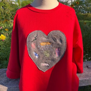 May include: A vibrant red sweatshirt with a large, grey heart-shaped appliqué. The heart is decorated with colourful, embroidered details. The sweatshirt has long sleeves with rolled cuffs and a classic crew neck. The garment is displayed on a mannequin.