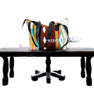 Puede incluir: Un bolso tote tejido con un patrón del suroeste en marrón, beige, turquesa y blanco. El bolso tiene asas y ribetes de cuero negro. El bolso está sobre una mesa de madera oscura.