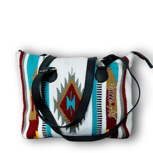 Puede incluir: Bolso tote blanco con un colorido patrón tejido en rojo, turquesa y dorado. El bolso presenta correas de cuero negro y el texto "Piazzo Art" en cursiva.