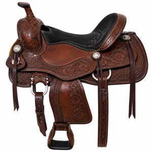 May include: A brown leather western saddle featuring intricate floral tooling, a smooth black padded seat, silver-toned conchos, and leather tie strings. The saddle includes a horn, stirrups, and adjustable straps with metal hardware and buckles.