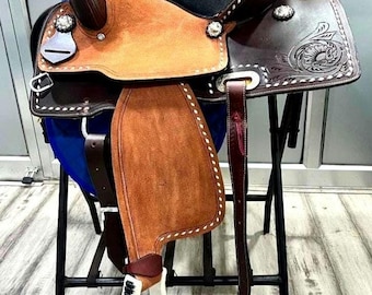 May include: A western saddle featuring a two-tone design with dark brown leather and lighter tan suede accents. The saddle includes intricate floral tooling on the skirts, silver-toned conchos, white stirrups with black tread, and a black seat.
