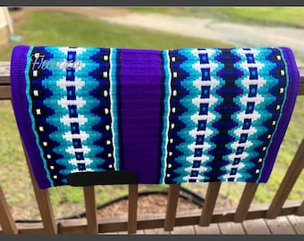 May include: A vibrant purple and turquoise western saddle pad with a geometric pattern. The pad features a central purple panel and a black leather patch. The design includes white and black accents, creating a striking visual contrast.