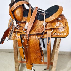 May include: A tan leather western saddle featuring intricate floral tooling, a black suede seat, and silver-toned hardware. The saddle includes a horn, stirrups, and leather straps, with the brand name FRAZZO printed in white on the side panel.