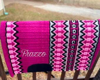 May include: A vibrant pink saddle pad with a decorative pattern in black, white, and pink. The pad features the name "Frazzo" embroidered in a cursive font. The pad is designed for equestrian use.