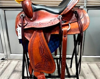 May include: A brown leather western saddle with intricate floral tooling. The saddle features a black seat, stirrups, and a yellow pad. The saddle is displayed on a black metal stand, showcasing its detailed craftsmanship and design.