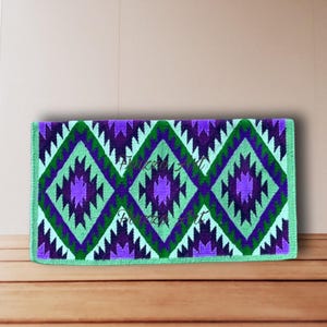 May include: A rectangular, woven textile featuring a repeating geometric pattern of purple, green, and white diamonds. The design is bordered by a light green edge. The textile is displayed on a wooden surface.
