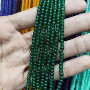 May include: Strands of small, round, dark green beads are held in a hand. The beads are strung together, creating a necklace or bracelet material. Other strands of colorful beads are visible in the background.