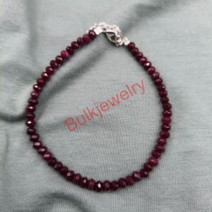 May include: A burgundy beaded bracelet with a silver clasp. The beads are small and faceted, reflecting light. The bracelet is displayed on a gray surface, with the text "Bulkjewelry" in the lower center.