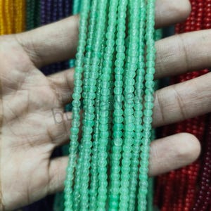 May include: Strands of small, round, light green beads. The beads are strung together, creating a delicate and textured appearance. The beads are held in a hand, with other strands of beads in the background.