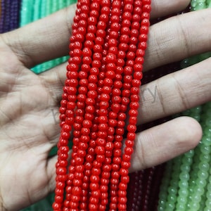 May include: Close-up of strands of small, round, bright red beads. The beads are strung together, creating a textured surface. The beads are held in a hand, with other strands of beads in the background.