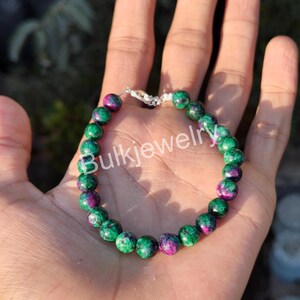 May include: A beaded bracelet featuring round, polished stones in vibrant shades of emerald green and deep magenta. The beads are strung together on a flexible cord and secured with a silver-toned metal clasp. The text Bulkjewelry is visible.