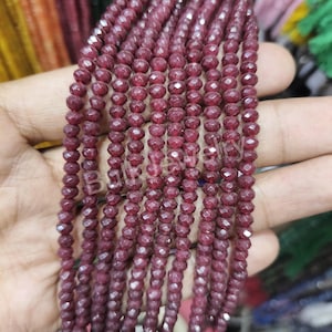 May include: Strands of faceted, deep red gemstone beads. The beads are small, round, and strung together, creating a textured surface. The color is a rich, dark red, with a slight sparkle from the facets. The beads are held in a hand.