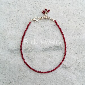 May include: A delicate red beaded bracelet with a silver clasp and a small cluster of red beads at the end. The bracelet is displayed on a gray surface. The text "Bulk Jewelry" is visible on the surface.