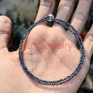 Puede incluir: Un delicado collar de doble hebra con pequeñas cuentas negras facetadas. El collar se sostiene en una mano, con el texto "Bulk Jewelry" visible en el fondo. Las cuentas parecen ser de tamaño uniforme y están ensartadas.