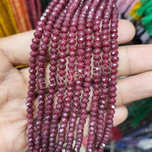 Puede incluir: Primer plano de varios hilos de cuentas de piedras preciosas de color rojo intenso. Las cuentas son pequeñas, redondas y facetadas, creando una superficie texturizada. El color es un rojo oscuro intenso, con un ligero brillo. Ideal para la creación de joyas.