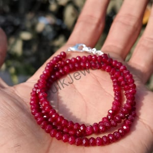 Può includere: Una collana di perline rosso intenso con chiusura argentata. La collana è composta da piccole perline sfaccettate, creando un effetto scintillante. Le perline sono di un ricco colore simile al rubino e la collana è esposta su una mano.