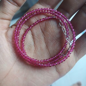 Könnte beinhalten: Eine zarte Halskette mit mehreren Strängen aus kleinen, facettierten, tiefrosa Edelsteinen. Die Halskette wird in einer Hand gehalten, die die leuchtende Farbe und das aufwendige Design zeigt. Der Verschluss ist silberfarben.