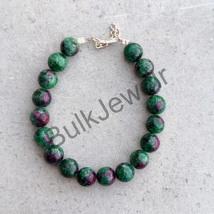 Könnte beinhalten: Ein Armband aus runden, polierten Perlen mit einem gesprenkelten Muster in Grün und tiefem Magenta. Das Armband wird mit einem silberfarbenen Karabinerverschluss aus Metall und einer kurzen Verlängerungskette geschlossen. Text BulkJewelry sichtbar.