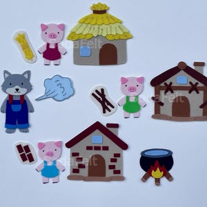 Peut inclure: Un ensemble de découpes en feutrine comprenant trois cochons vêtus de tenues colorées, un loup gris en salopette bleue, trois maisons distinctes en paille, bois et briques, une botte de paille, un nuage de souffle, des bâtons, des briques et un chaudron.