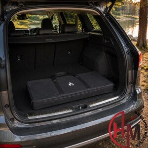 May include: A rectangular black carrying case with a white flame logo on the front, positioned inside the open trunk of a dark gray vehicle. The case features two black fabric handles and a zippered closure, resting flat on the dark interior floor.