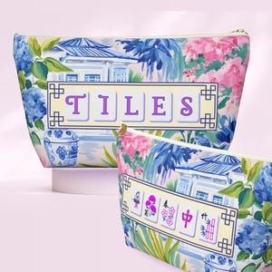 Op de afbeelding: Twee cosmetische tassen met een bloemen- en architectonisch ontwerp. De tassen hebben een lichte achtergrond met blauwe en roze bloemen, een blauw en wit gebouw en het woord "TILES" in paarse letters. De ene tas is groter dan de andere.